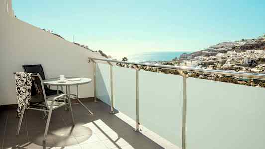 Altan med bord og stol, udsigt over hav og bygninger. (AI-genereret tekst)