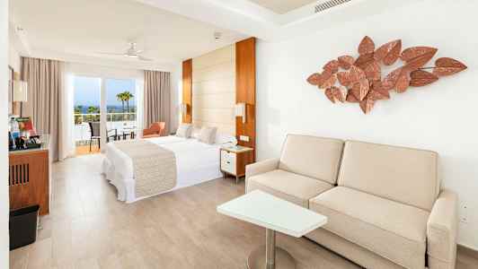 Lyst hotelværelse med seng, sofa og balkon samt havudsigt. (AI-genereret tekst)