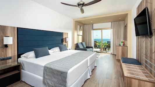 Et hotelværelse med to senge, et siddeområde og en balkon med udsigt over havet. (AI-genereret tekst)