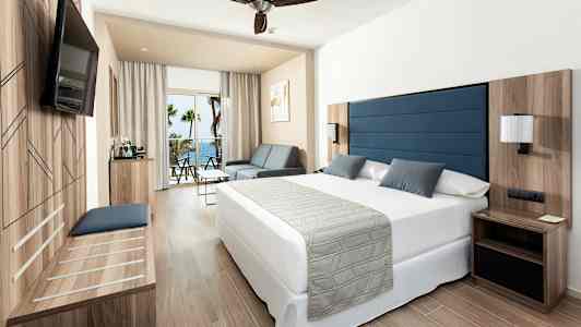 Et hotelværelse med en stor seng, en sofa og et bord. Der er en balkon med udsigt over havet. (AI-genereret tekst)