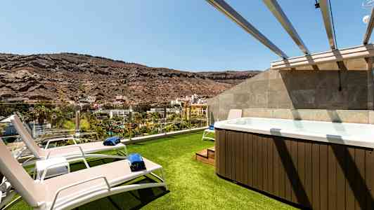 Billede af en terrasse med græsbelagt område, liggestole og et boblebad med udsigt over et bjergrigt landskab. (AI-genereret tekst)