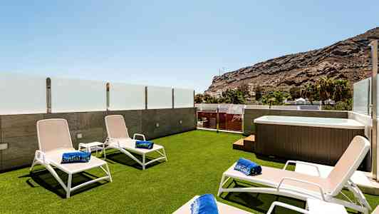 Terrasse med liggestole, badehåndklæder og jacuzzi, omgivet af glasværn og udsigt til bjergene. (AI-genereret tekst)