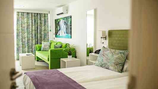 Værelse med seng og grøn sofa, med farverige puder og gardiner samt kunst på væggen. (AI-genereret tekst)