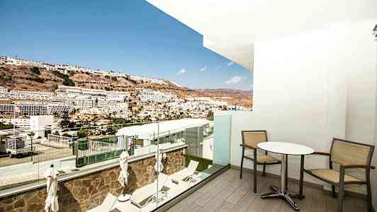 Udsigt fra en altan med bord og stole ud mod en skråning med bygninger i det fjerne. (AI-genereret tekst)