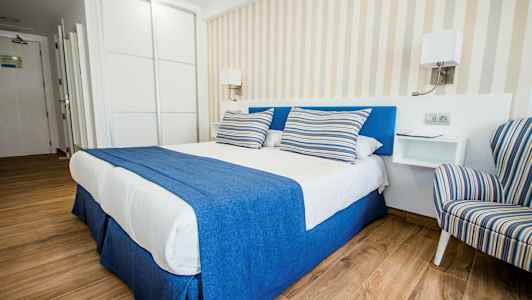 Et stilfuldt hotelværelse med en dobbeltseng med blåt og hvidt sengetøj samt stribede puder, ved siden af en stribet lænestol. (AI-genereret tekst)