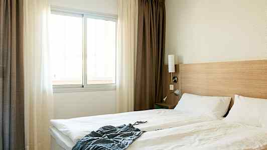 Et hotelværelse med en dobbeltseng, hvide sengelagner, en trævæg ved sengegavlen og et vindue med lyse gardiner. (AI-genereret tekst)