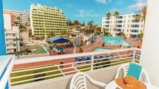 Udsigt fra en balkon, der viser et hotelområde med pool og liggestole samt bygninger i baggrunden. (AI-genereret tekst)