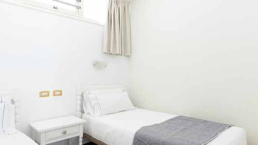 To enkeltsenge med hvidt sengetøj og et gråt tæppe, placeret i et lyst rum. (AI-genereret tekst)