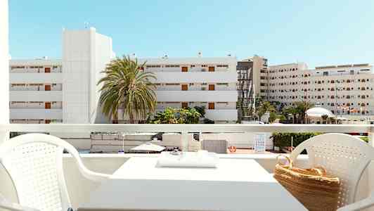 Udsigt fra en altan med siddegruppe, bygninger og palmer synlige i baggrunden under en klar blå himmel. (AI-genereret tekst)