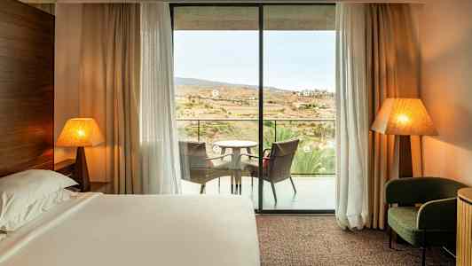 Et hotelværelse med en seng, to bordlamper, en lænestol og en altan med siddepladser. (AI-genereret tekst)