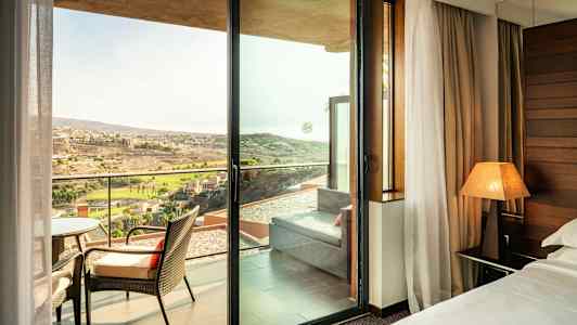 Et hotelværelse med udsigt gennem en glasdør til en altan med stole og et fjernt landskab udenfor. (AI-genereret tekst)