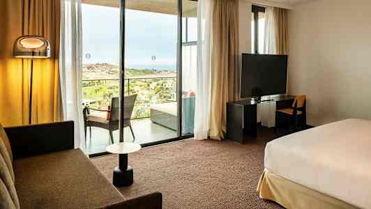 Hotelværelse med siddeområde, seng, TV og udsigt til en balkon med landskab udenfor. (AI-genereret tekst)