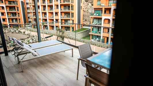 To liggestole og et bord med stole på en rummelig altan med udsigt til en gårdhave samt flere orange bygninger. (AI-genereret tekst)