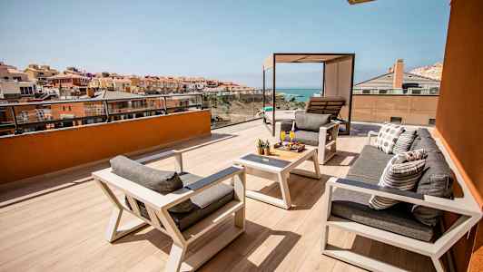 En rummelig terrasse med siddearrangement og borde, udsigt til havet og omkringliggende bygninger. (AI-genereret tekst)