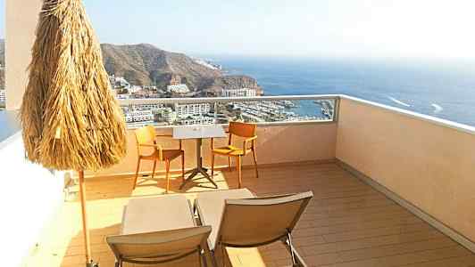 Udsigt fra en terrasse med parasol, to stole og et bord samt liggestole med havudsigt. (AI-genereret tekst)