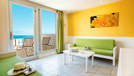 En lys stue med en grøn sofa, et hvidt sofabord og adgang til en altan med udsigt over havet. (AI-genereret tekst)