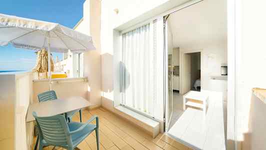 En altan med bord og stole, parasol og udsigt mod havet. En dør leder ind til et lyst interiør med møbler. (AI-genereret tekst)