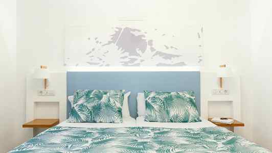 Seng med grønt og hvidt mønstret tæppe og puder, med blå sengegavl og hvide vægge. (AI-genereret tekst)