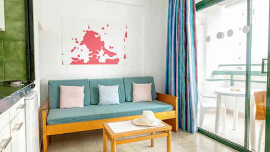 En stue med en turkis sofa, farverige puder, bord og en hat. Et vindue med udsigt til en altan. (AI-genereret tekst)