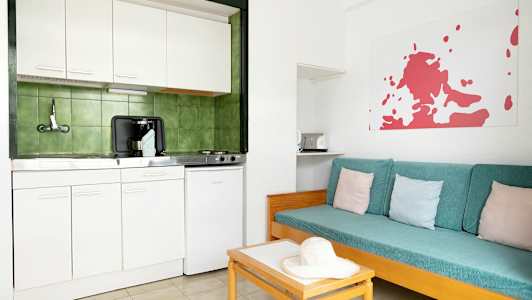 Et enkelt køkken med hvidt interiør og grønne fliser bagved. Ved siden er en sofa med turkise puder og pastelfarvede pudebetræk. (AI-genereret tekst)