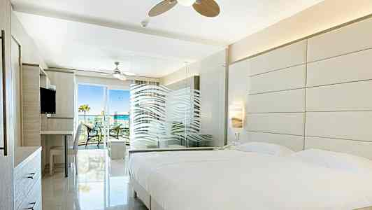 Lyst hotelværelse med stor seng, glasvæg og adgang til altan. (AI-genereret tekst)