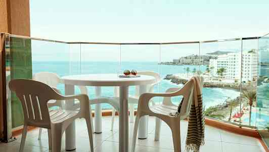 Udsigt fra en altan med et bord og plastikstole, havet og bygninger synlige i baggrunden. (AI-genereret tekst)