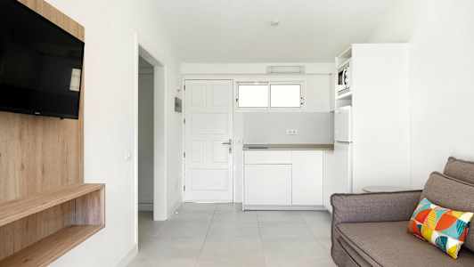 Et hvidt rum med en sofa, tv, træhylde og køkken. (AI-genereret tekst)