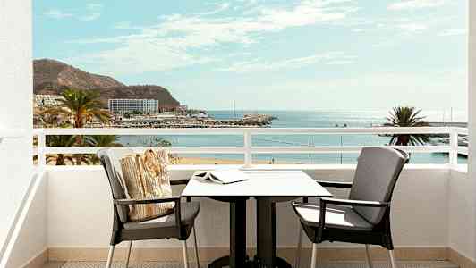 Udsigt fra en altan med bord og stole mod en strand og havet. (AI-genereret tekst)