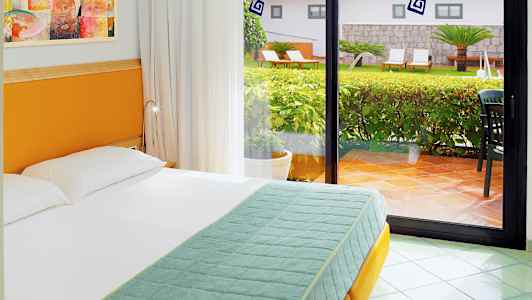 Et hotelværelse med en seng foran et stort glasvindue med udsigt til en frodig have. (AI-genereret tekst)