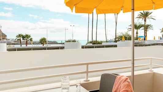 En altan med et bord, et orange håndklæde på en stol, en karaffel og et glas på bordet samt gule parasoller og palmer i baggrunden. (AI-genereret tekst)