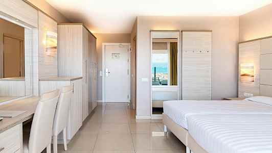 Et hotelværelse med moderne indretning, en dobbeltseng, garderobe og skrivebord, med udsigt til havet. (AI-genereret tekst)