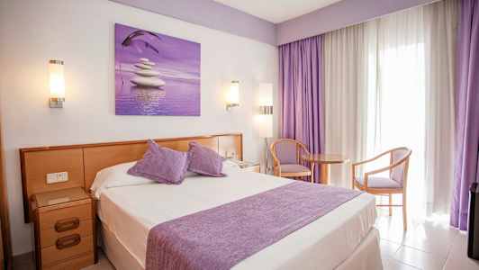 Hotelværelse med dobbeltseng, lilla detaljer og siddeplads ved et vindue. (AI-genereret tekst)