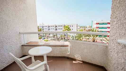 Billede af en altan med et bord og en stol, med udsigt over bygninger og palmer. (AI-genereret tekst)