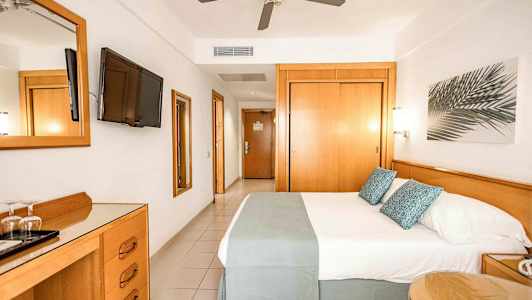 Et hotelværelse med en dobbeltseng, tv på væggen, loftventilator og træmøbler. (AI-genereret tekst)