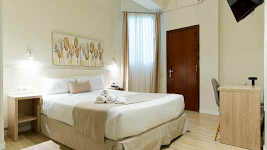 Et lyst hotelværelse med en dobbeltseng, et skrivebord og et vægmonteret TV. (AI-genereret tekst)