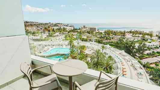 Altan med to stole og et rundt bord samt udsigt over hotelområde med pool og hav. (AI-genereret tekst)