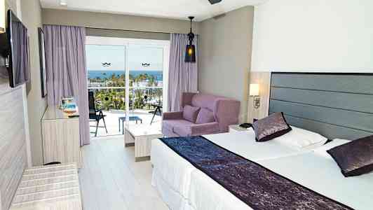 Moderne hotelværelse med en dobbeltseng, sofagruppe og altan med udsigt over havet. (AI-genereret tekst)