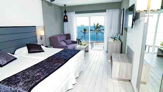 Hotelværelse med dobbeltseng, sofa og havudsigt uden for store vinduer. (AI-genereret tekst)