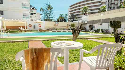 En udendørs pool omgivet af grønne områder og liggestole, med hvide bygninger i baggrunden. (AI-genereret tekst)