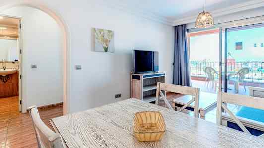 Et opholdsområde med et bord, stole og en tv, samt en altan i baggrunden. (AI-genereret tekst)