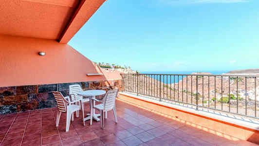 Terrasse med bord, stole og rækværk, samt havudsigt. (AI-genereret tekst)