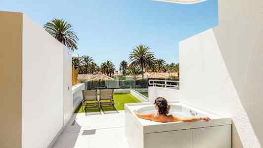 En person sidder i et boblebad på en terrasse med udsigt over palmer og huse. (AI-genereret tekst)