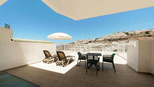 En terrasse med liggestole, parasol og havemøbler med udsigt mod solen og en bjergskråning. (AI-genereret tekst)