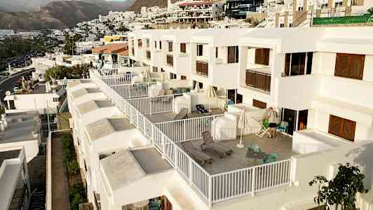 Billede af en række moderne hvide lejligheder med store private altaner, møbleret med stole og liggestole, i et kuperet landskab med bjerge i baggrunden. (AI-genereret tekst)