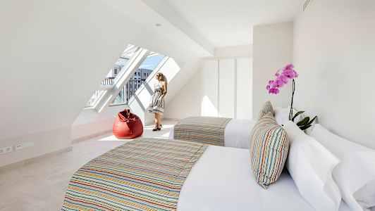 Et lyst værelse med to senge, farverige puder, en rød sækkestol og et stort skråstillet vindue. (AI-genereret tekst)