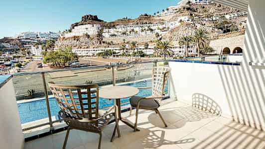 En balkon med to stole og et bord, udsigt over en pool, bygninger og bjerge. (AI-genereret tekst)