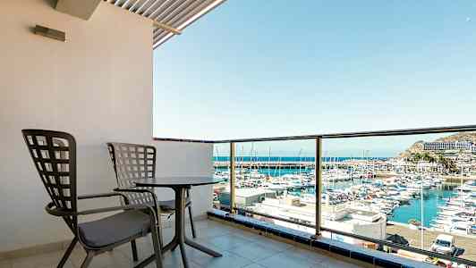 Billedet viser en altan med to stole og et bord, med udsigt over en havn med både. (AI-genereret tekst)