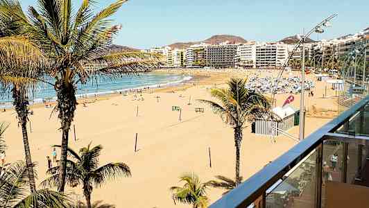 Udsigt over en bred sandstrand med palmer og byens bygninger i baggrunden. (AI-genereret tekst)