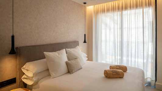 Et hotelværelse med en stor seng, hvide lagner og puder. På sengen ligger sammenrullede håndklæder. Vægge og gardiner er lyse, og rummet har hængende lamper. (AI-genereret tekst)