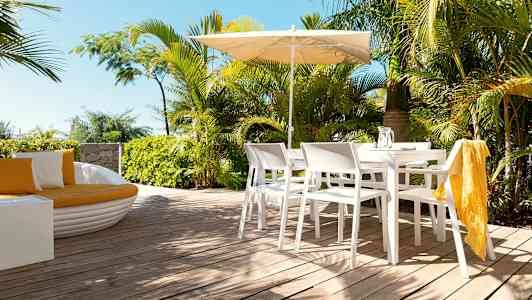 En terrasse med hvide stole og bord under en parasol, omgivet af grønne planter. (AI-genereret tekst)